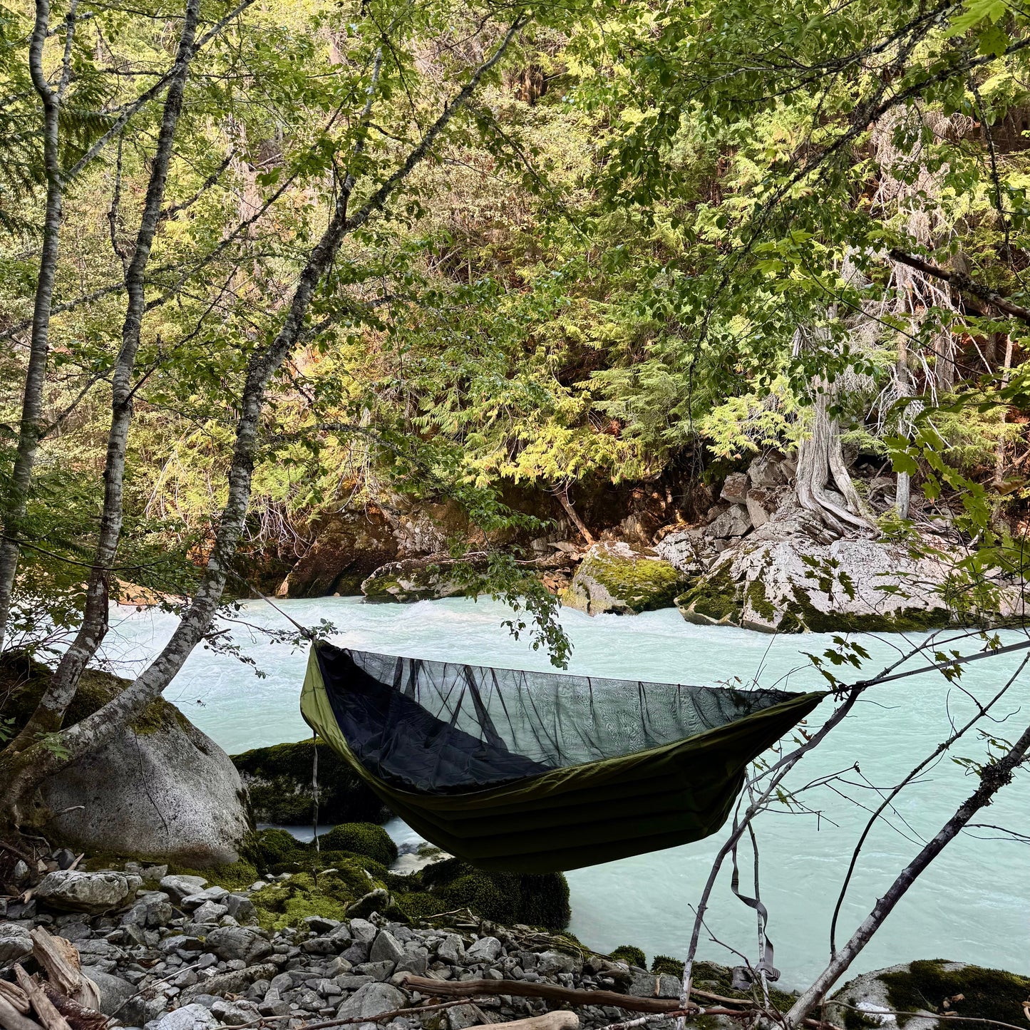 Backpacking Hammock hanging between trees over a glacier blue river in a forest setting