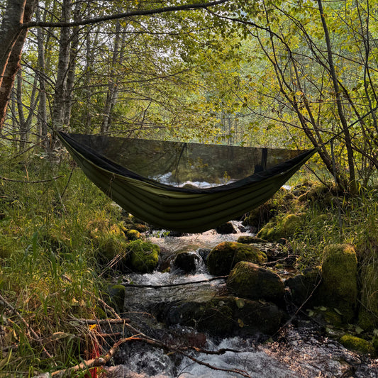 Insulated backpacking hammock hanging between trees in a forest with a stream below